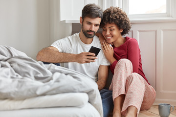Friendly multiethnic woman, man read some text or positive news in internet on mobile phone, dressed in casual domestic clothes, sit on floor near bed, connected to wireless internet, recreation time