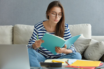 Beautiful Caucasian woman in striped sweater and spectacles, concntrated on homework, poses at comfortable couch in modern apartment, uses laptop computer for chatting online, poses at home.
