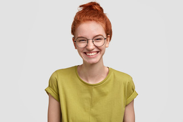 Positive freckled pupil has red hair, smiles broadly, wears round optical glasses and green t shirt, stands against white background, rejoices good moments in school life, remembers something pleasant