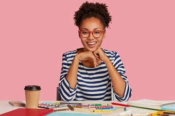 Fashion designer with toothy smile, feels sarisfied, keeps hands under chin, wears striped clothes, isolated over pink background. Positive painter has inspiration for drawing. Creative work concept