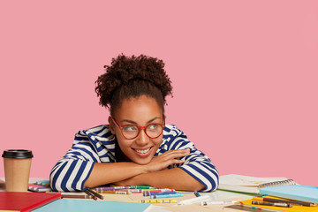 Creativity and inspiration concept. Pleased smiling Afro American graphic designer leans at table, has coffee break after drawing picture in notebook, isolated over pink background in art studio