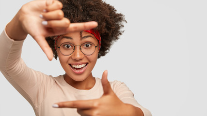 Smiling delighted female designer makes frame gesture with fingers, stands over white wall,...