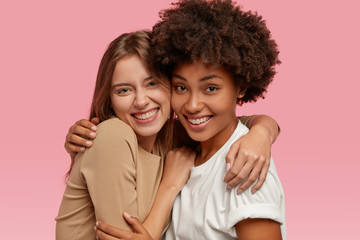 Photo of cheerful friendly interracial females have warm embrace, smile gladfully and look at camera, pose for family portrait, dressed in casual clothes, isolated over pink background. Friendship