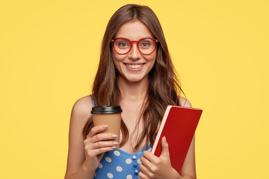 Photo of cheerful female college student carries execise book and take out coffee, smiles broadly, being in good mood after lectures, rejoices coming holidays, models against yellow studio wall