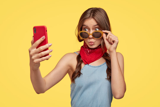 Isolated shot of fashionable teenage girl pouts lips, looks through trendy sunglasses, wears red bandana near neck, holds smart phone, makes selfie portrait, enjoys spare time, stands over yellow wall