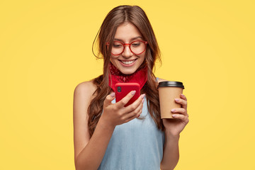 Cheerful girl has coffee break, rejoices buying new gadget, reads notification on red mobile phone,...