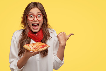 Pleased happy young woman gazes with happiness, points with thumb aside at free space, eats pizza,...