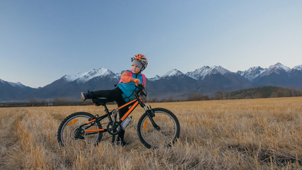 Fototapeta premium One caucasian children walk with bike in wheat field. Little girl walking black orange cycle on background of beautiful snowy mountains. Biker stand with backpack and helmet. Mountain bike hardtail.