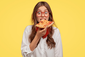 Emotive beautiful lady bites delicious pizza, looks directly at camera, has time for snack, visits...