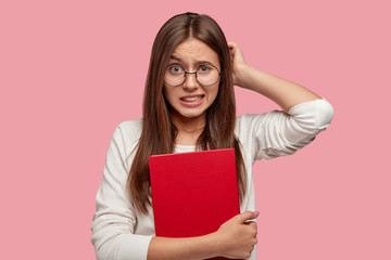 College student scratches head in bewilderment, clenches white teeth, remembers information before answering on final exam, holds red exercise book, feels displeased, has problems with studying.