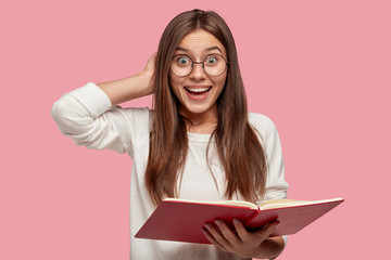 Pretty smiling girl has joyful expression, keeps hand behind head, carries red textbook, smiles broadly as reads necessary information, isolated over pink background, wears round spectacles.