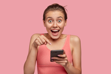 Photo of emotive Caucasian sporty woman has surprised expression, indicates with index finger in monitor of cellular, wears casual vest, recieves good notification, isolated over pink background
