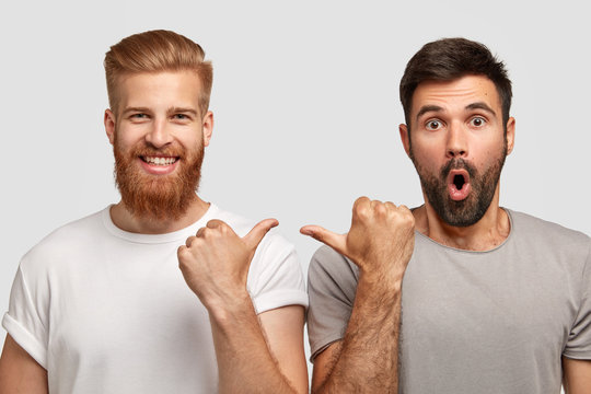 Shocked Unshaven Man With Dark Stubble Indicates At His Companion, Wears Grey T Shirt. Cheerful Ginger Guy With Trendy Haircut And Bristle Points At Fellow. Two Friends Model Indoor Over White Wall