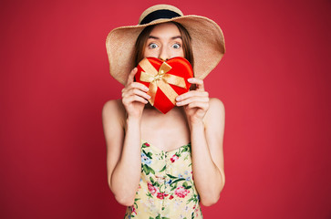 Beautiful surprised smiling blond woman in hat and dress holds gift box in shape of heart. Valentine present day