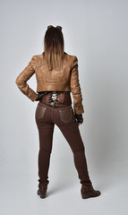 full length portrait of brunette  girl wearing brown leather steampunk outfit. standing pose with back to the camera,  on grey studio background.