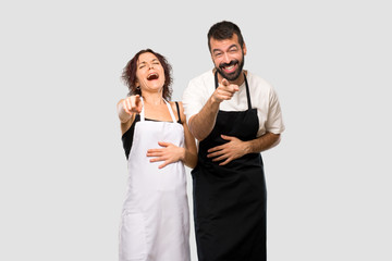 Couple of cooks pointing with finger at someone and laughing a lot on grey background