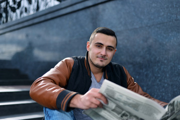 portrait of young guy in leather jacket with bag smiling and reading newspaper