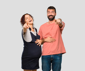 Couple with pregnant woman pointing with finger at someone and laughing a lot on isolated grey background