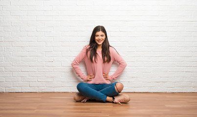 Teenager girl sitting on the floor in a room posing with arms at hip and laughing looking to the front