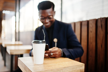 Amazingly looking african american man wear at blue blazer with brooch, black turtleneck and glasses posed at street. Fashionable black guy with cup of coffe looking at his phone.
