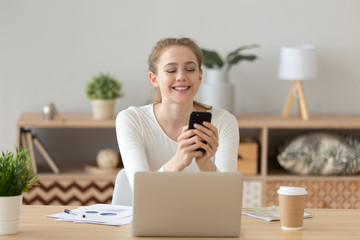 Obraz premium Happy millennial female sit at table with laptop reading pleasant message on smartphone, smiling young woman chatting on cellphone, texting with boyfriend or friend, excited girl get good news on cell