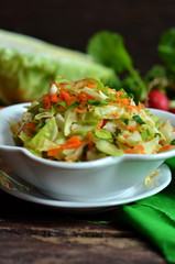 Young cabbage salad with carrot in a white bowl on a wooden background. Radishes and young cabbage in the background