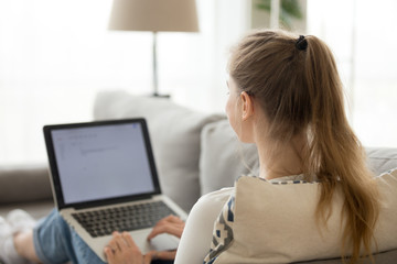 Girl sitting on cozy couch with laptop on knees browsing internet or checking email, female relax...