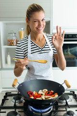 Portrait of healthy young woman looking at camera while cooking and mixing food in frying pan in the kitchen at home.