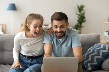 Excited husband and wife sit on couch win lottery online at home, happy millennial couple look at...