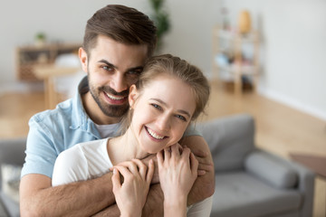 Fototapeta premium Portrait of happy millennial couple hug looking at camera, smiling husband hold in arms beloved young wife posing in living room, headshot of excited first time buyers make family picture in new home