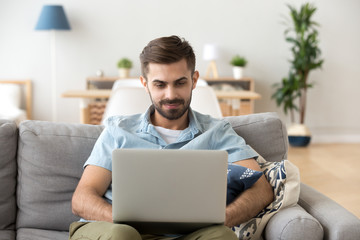 Focused millennial man sit on couch in apartment using laptop browsing internet, young male relax...