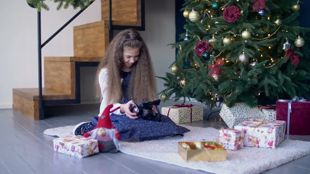 Cheerful preadolescent girl viewing photos on digital camera while sitting under the christmas tree, surrounded by gift boxes. Smiling cute child looking through pictures on photo camera at xmas.