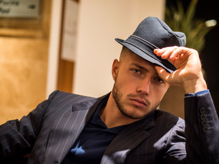 Handsome elegant man in hotel or night club lounge, wearing business suit and fedora hat, looking at camera - obrazy, fototapety, plakaty