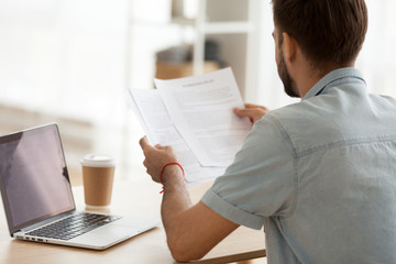 Back view of focused man sitting at office table working at laptop looking at paper documents, concentrated male holding paperwork considering issue, busy using computer and drinking coffee