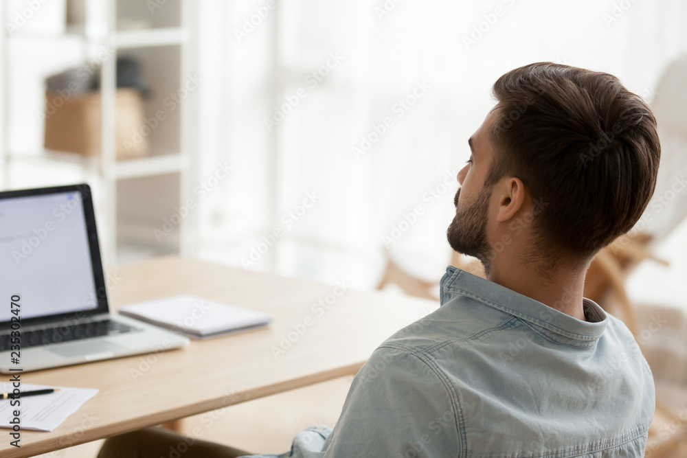 Wall mural Back view of exhausted millennial man sit at workplace falling asleep, tired male worker distracted from work taking break or nap near laptop, sleepy guy relax with eyes closed sleeping near computer