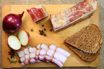 Smoked bacon, sliced on a wooden plank. Smoked bacon, red onion, salt and pepper on a clay dish. Smoked lard.