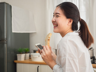 asian woman eating bread while using smart phone in the kitchen