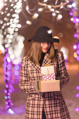 Young girl standing in front on christmas tree lights holding presents and gifts and smiling at winter alone in the park
