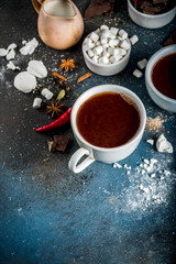 Obraz na płótnie Canvas Hot chocolate cups, broken pieces of chocolate, sugar, marshmallow, red chili peppers and spices. Dark blue concrete background top view space