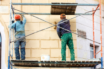 Naklejka na ściany i meble two workers in a helmet and construction clothes on a high staircase are painting with rollers, repairing the facade of the house