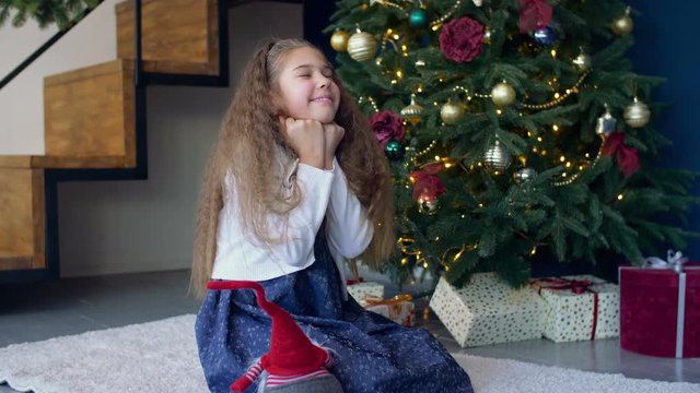Positive joyful little girl with eyes closed tightly, propped her head on clenched fists making a wish, asking Santa Claus to fulfill dreams at christmas time over festive decorated room background.