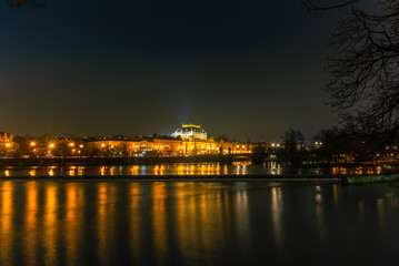 Fototapeta na wymiar Illuminated classical buildings and street lights reflecting on the Vltava river in Prague before a small waterfall in a cold winter night - 1