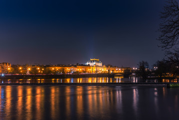 Fototapeta na wymiar Illuminated classical buildings and street lights reflecting on the Vltava river in Prague before a small waterfall in a cold winter night - 2