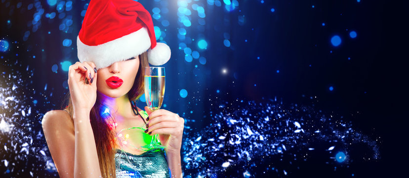 Christmas Sexy Woman. Beauty Model Girl In Santa's Hat With Glass Of Champagne In Her Hand Celebrating On Blinking Holiday Winter Wide Background