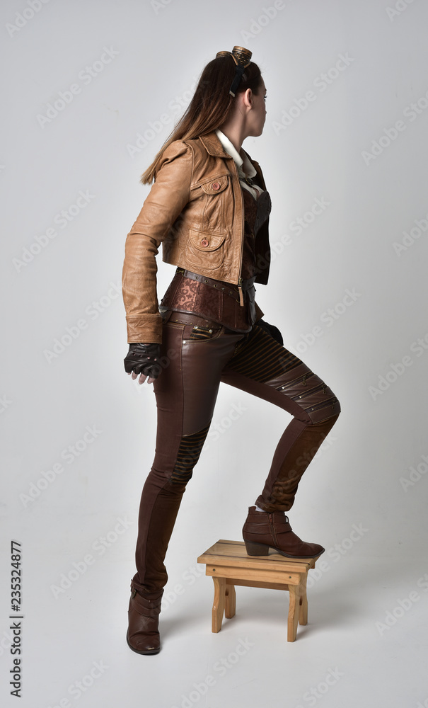 Canvas Prints full length portrait of brunette  girl wearing brown leather steampunk outfit. standing pose with back to the camera,  on grey studio background.