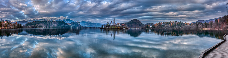 Fototapeta na wymiar Panoramic shot of the Lake Bled in Slovenia during twilight with the reflection of the surrounding hills and mountains