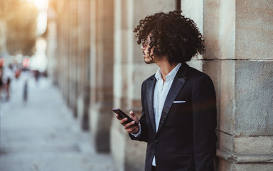 A young Asian man in a formal suit, sunglasses and with curly afro hair is looking aside while leaning against a stone column outdoors and typing a message on his smartphone