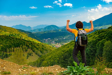 Naklejka na ściany i meble Young mountain hiker with arms raised up, celelbrating and enjoying a beautiful mountain landscape covered with lush forests. Hiking in a sunny summer day.