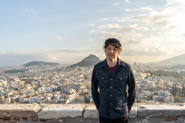 Portrait of a forty something man standing atop the Acropolis, and wearing a Greek sailor hat, with a wide view of the Athens, Greece cityscape beyond.