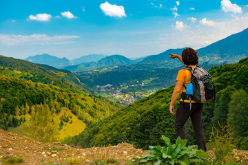 Young mountain hiker pointing to a beautiful mountain landscape covered with lush forests. Hiking in a sunny summer day.